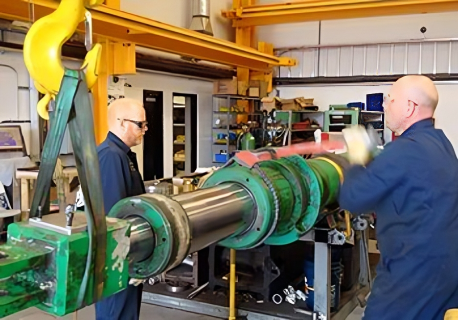 Two technicians rebuilding a large hydraulic cylinder lifted on a shop crane